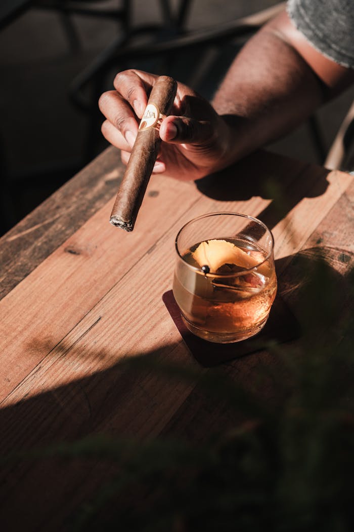A sophisticated setting featuring a hand holding a cigar and a cocktail glass on a rustic table, casting warm shadows.